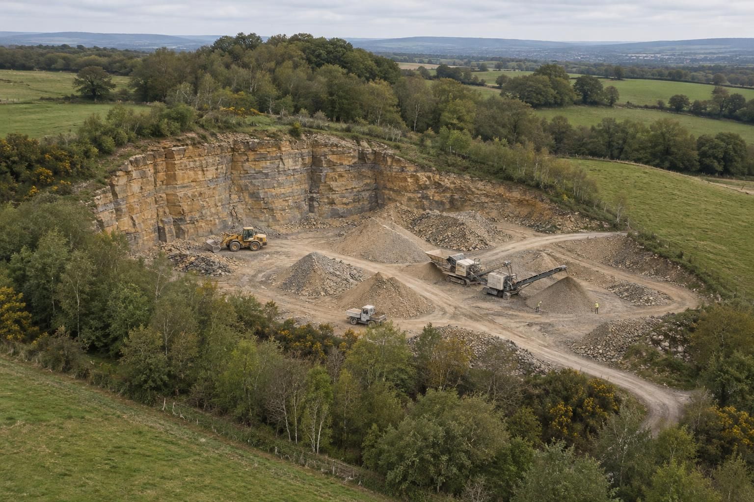 A high oblique aerial view of a small sandstone quarry set into green farmland: a compact worked area with buff faces and a few neat stockpiles, surrounded by thick woodland and hedged fields, a restored grassy slope along one edge, soft overcast light.