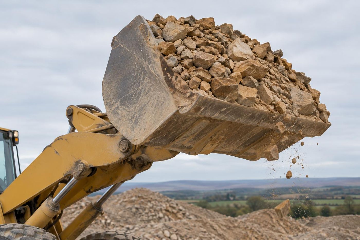 Close view of a loading shovel bucket heaped with crushed buff sandstone, lifted high, stone spilling from the lip, blurred stockpile behind.