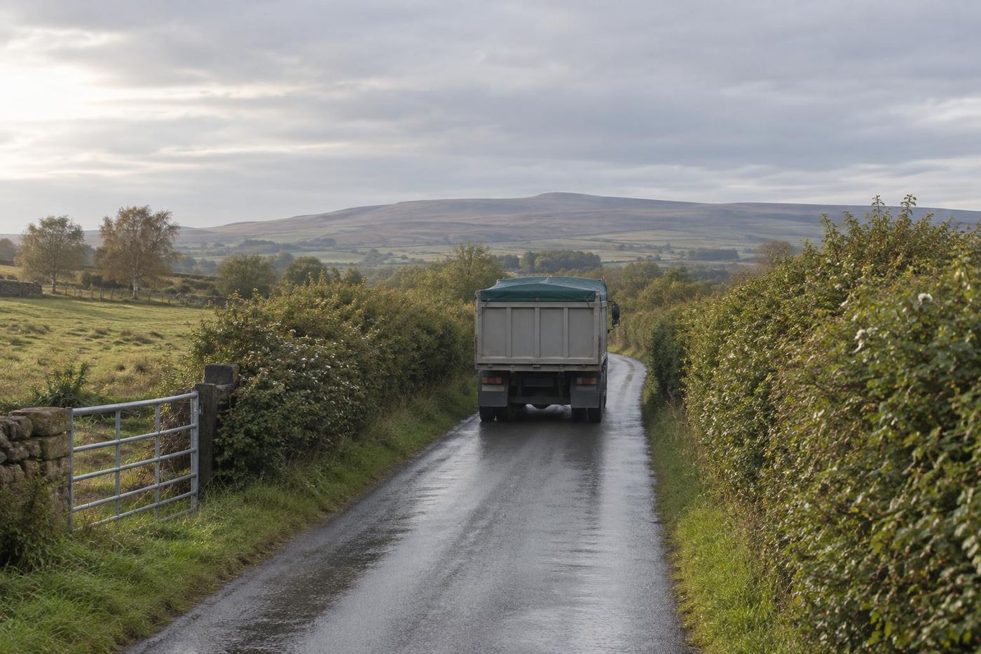 A sheeted plain grey tipper lorry leaving a quarry along a clean, swept tarmac access track between hedges, wet road with no mud, soft morning light, seen from behind.