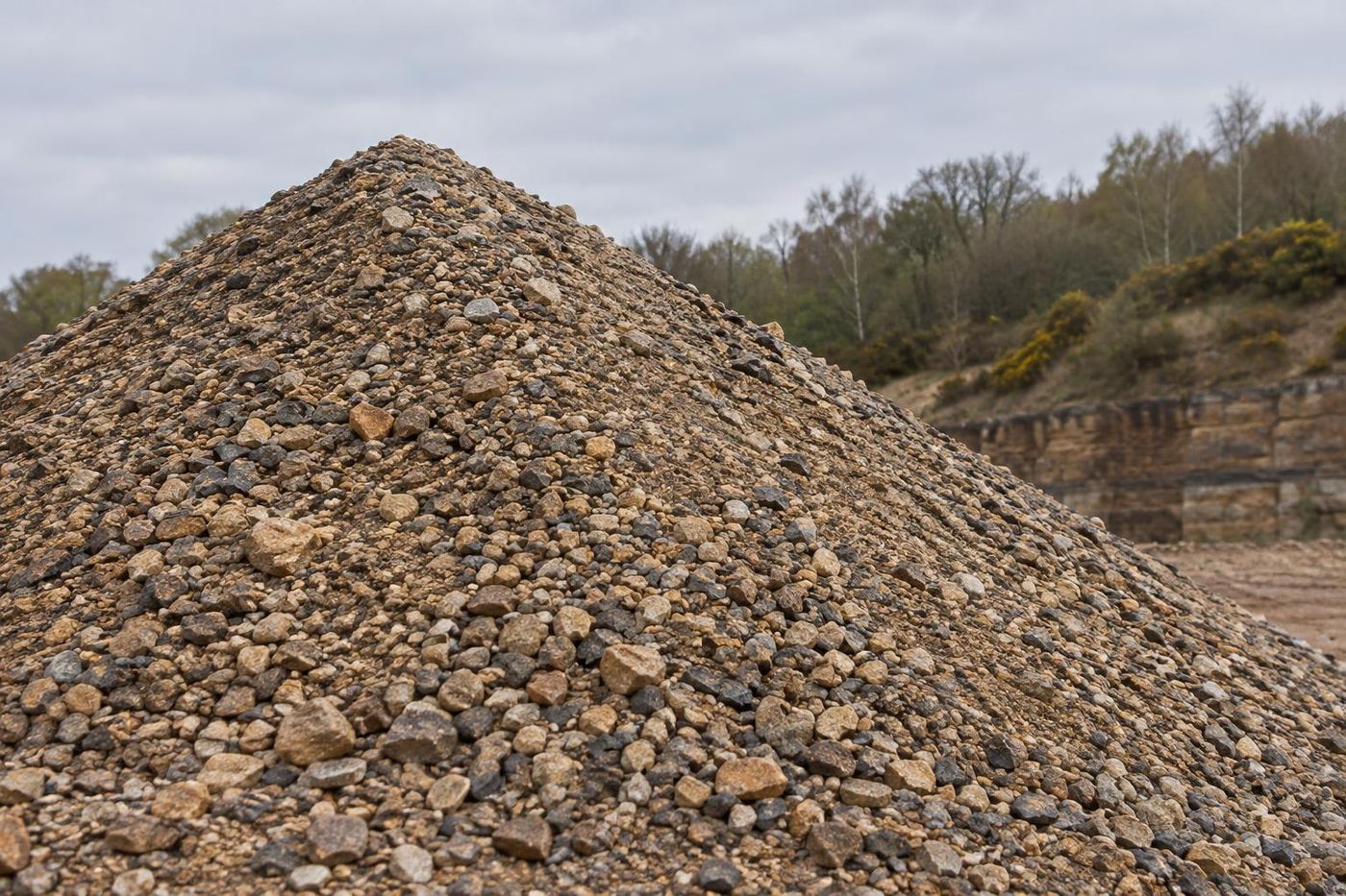 A tall conical stockpile of crushed sandstone sub-base with a visible mix of sizes from dust to coarse, damp surface, overcast light, wooded bank behind.
