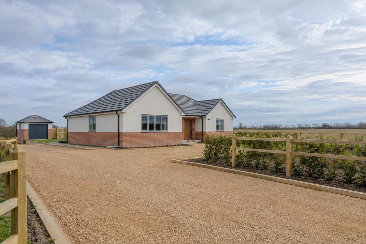 A newly built brick-and-render bungalow on a flat plot with a freshly rolled crushed sandstone sub-base drive in front, big flat-country sky.